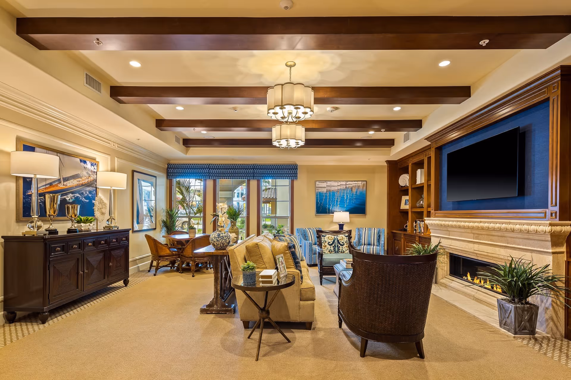 A cozy and elegant living room area with beige carpet, a beige sofa, two dark wicker chairs, and two blue striped armchairs. The room features a fireplace with a mounted flat-screen TV above it, wooden built-in shelves, and a sideboard with decorative lamps and trophies. Large windows with blue valances let in natural light, and the ceiling has wooden beams with two hanging light fixtures.