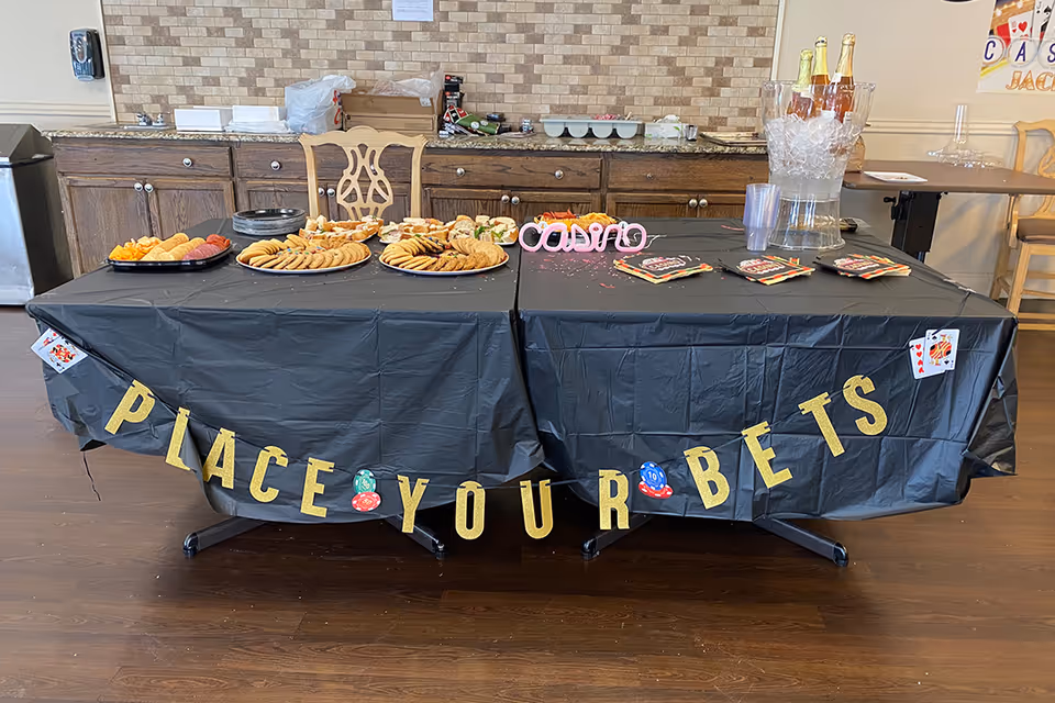 A table covered with a black tablecloth decorated with gold letters spelling 'PLACE YOUR BETS' and poker chip and playing card decorations. The table holds trays of assorted snacks including crackers, cookies, and sandwiches, as well as a clear ice bucket with bottles and plastic cups. The background shows a kitchen counter with cabinets and a brick-patterned backsplash.