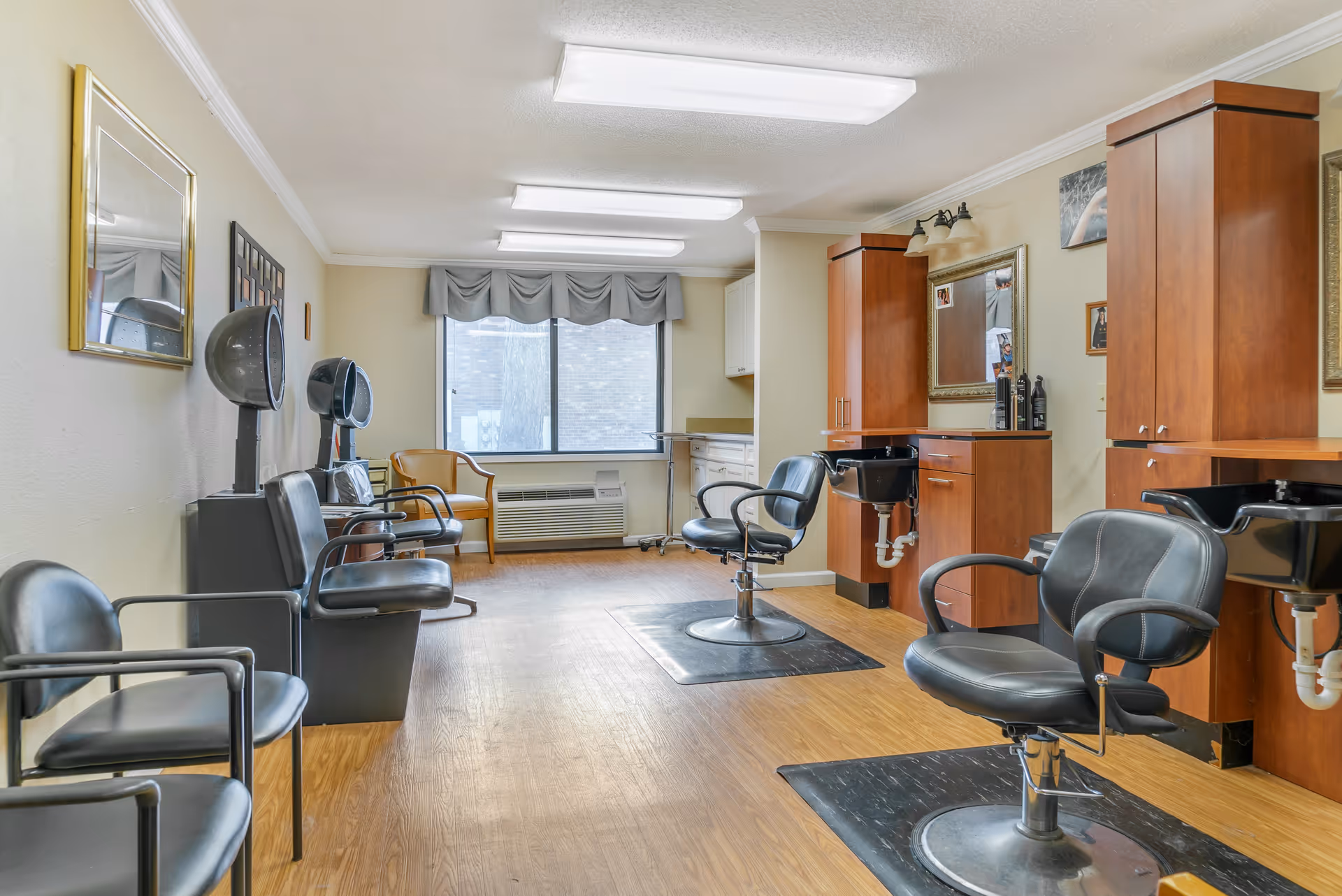 Interior of a hair salon area in a senior living facility with black salon chairs, hair drying stations, wooden cabinets with sinks, a large window with gray valance, and a wooden floor.