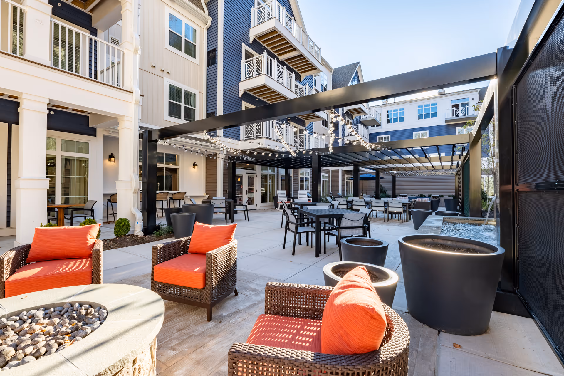 Outdoor patio area at a senior living facility with wicker chairs featuring orange cushions around a fire pit, multiple tables and chairs under a pergola with string lights, and a multi-story building with balconies in the background.