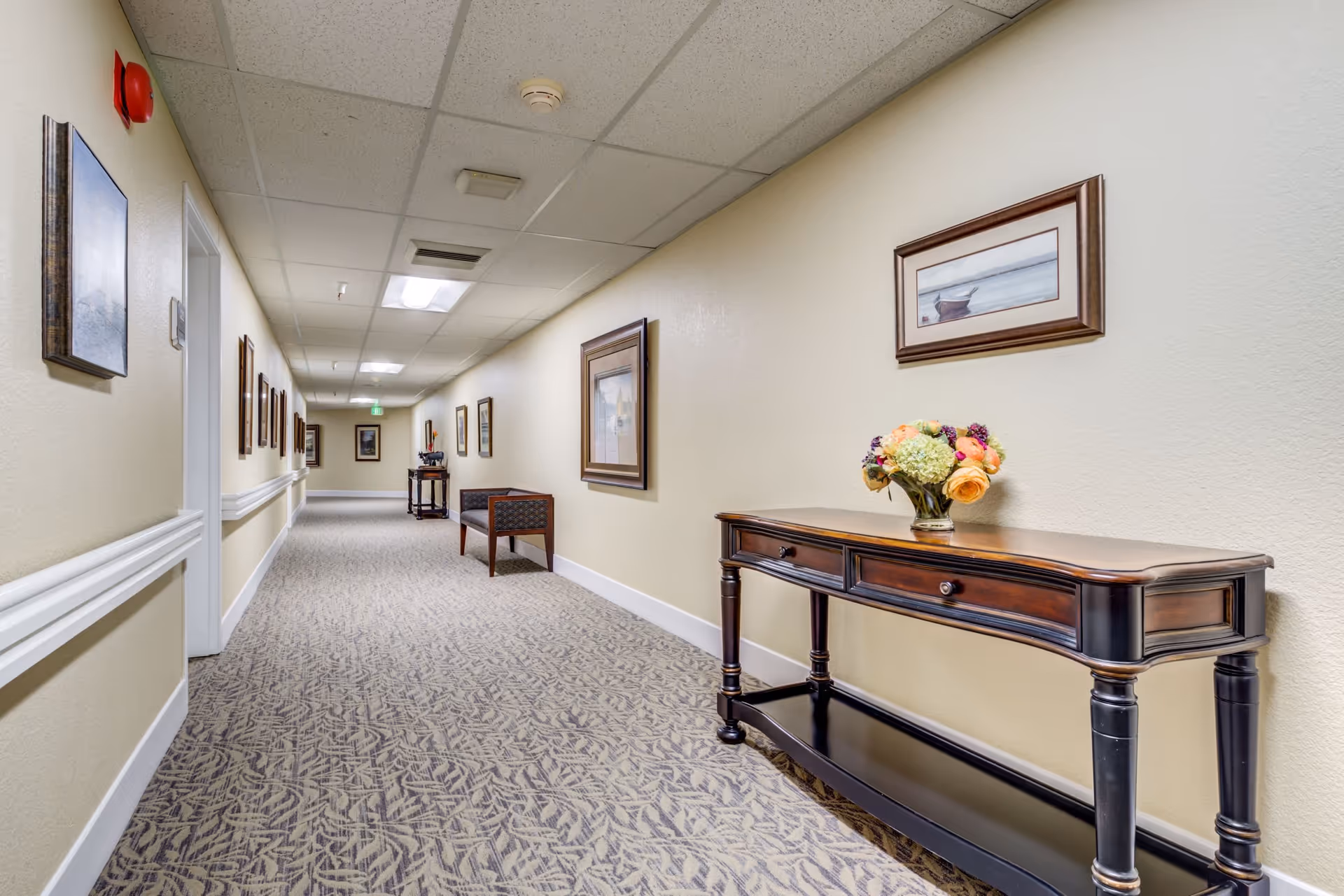 A well-lit hallway in a retirement community with beige walls and patterned carpet. The hallway features framed artwork on the walls, a wooden console table with a floral arrangement, and a bench further down the corridor. The ceiling has recessed lighting and a smoke detector.