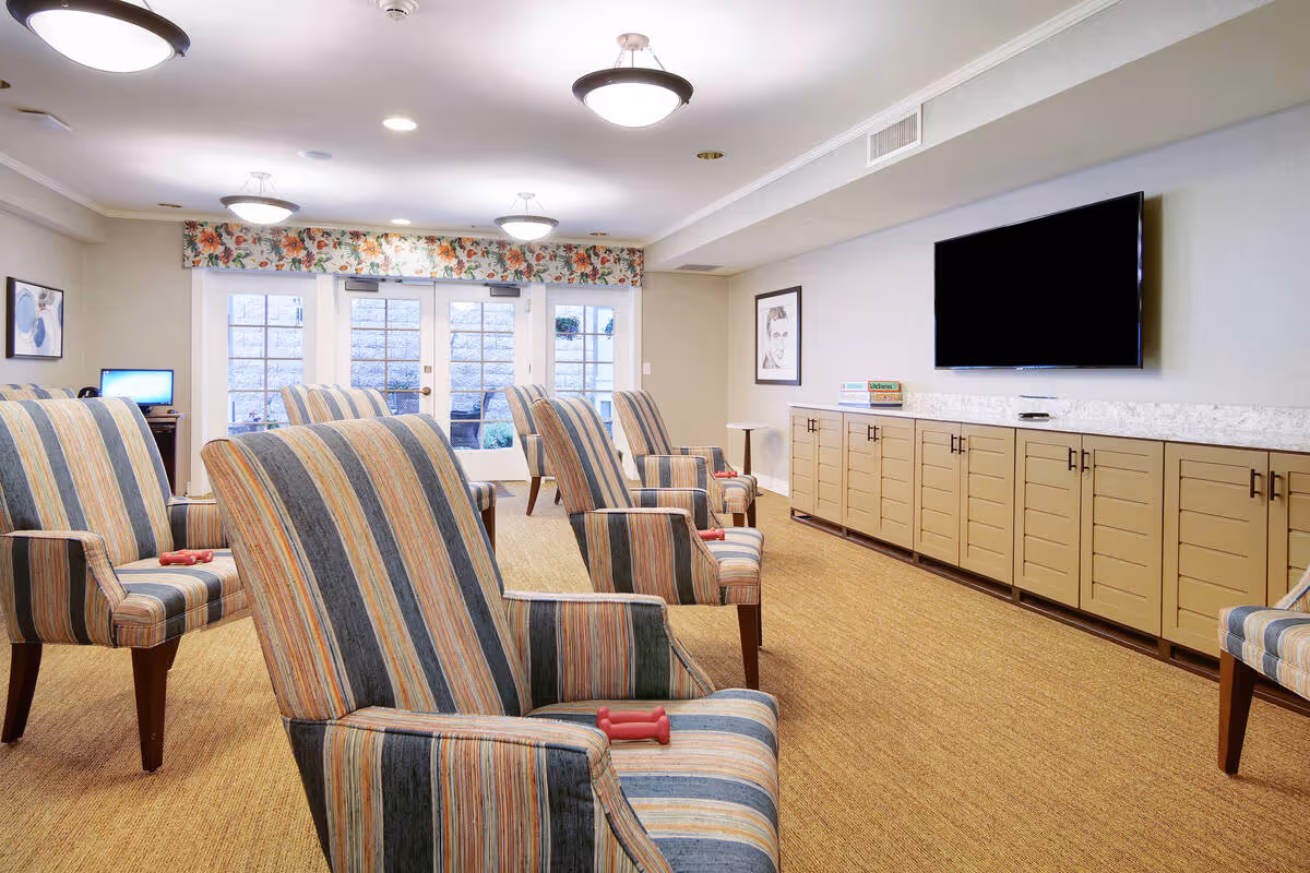 Comfortable common room with striped armchairs facing a wall-mounted TV and long cabinets, with windows and double doors at the far end.