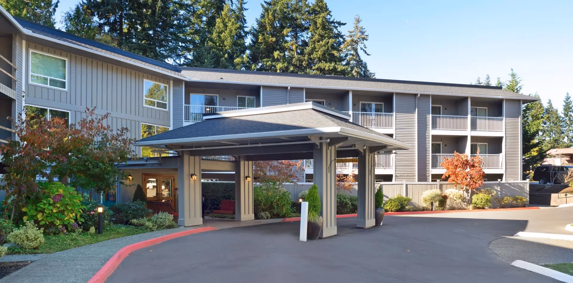 Exterior view of Aegis of Edmonds - Assisted Living facility showing a covered entrance with columns, surrounded by landscaped greenery and trees, with a multi-story building in the background featuring balconies and large windows.