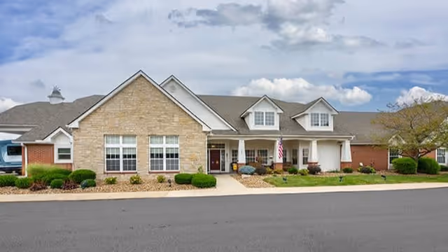 Front exterior of a one-story brick and stone senior living building with a covered entrance, American flag, and landscaped beds.