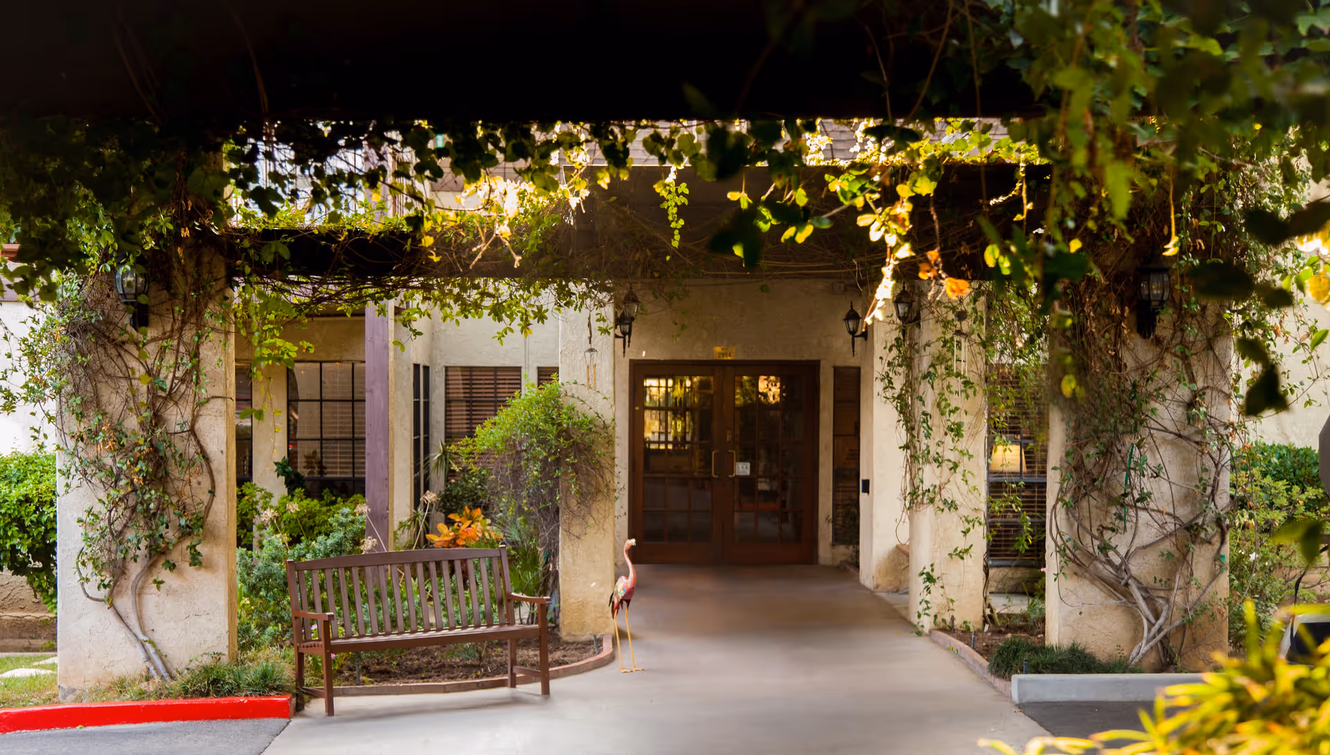 Entrance to a building with a wooden bench on the left side and a decorative flamingo statue next to it. The entrance is framed by columns covered with climbing plants and vines, with greenery surrounding the area and sunlight filtering through the foliage.