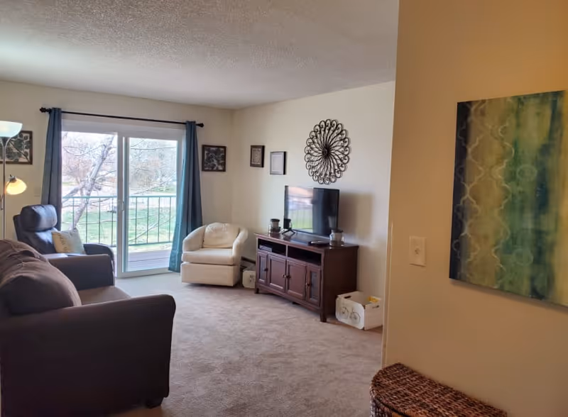 A cozy living room with a brown sofa, a white armchair, a dark wooden TV stand with a flat-screen TV, and wall decorations including framed pictures and a decorative metal wall piece. There is a sliding glass door with blue curtains leading to an outdoor area with trees visible outside.