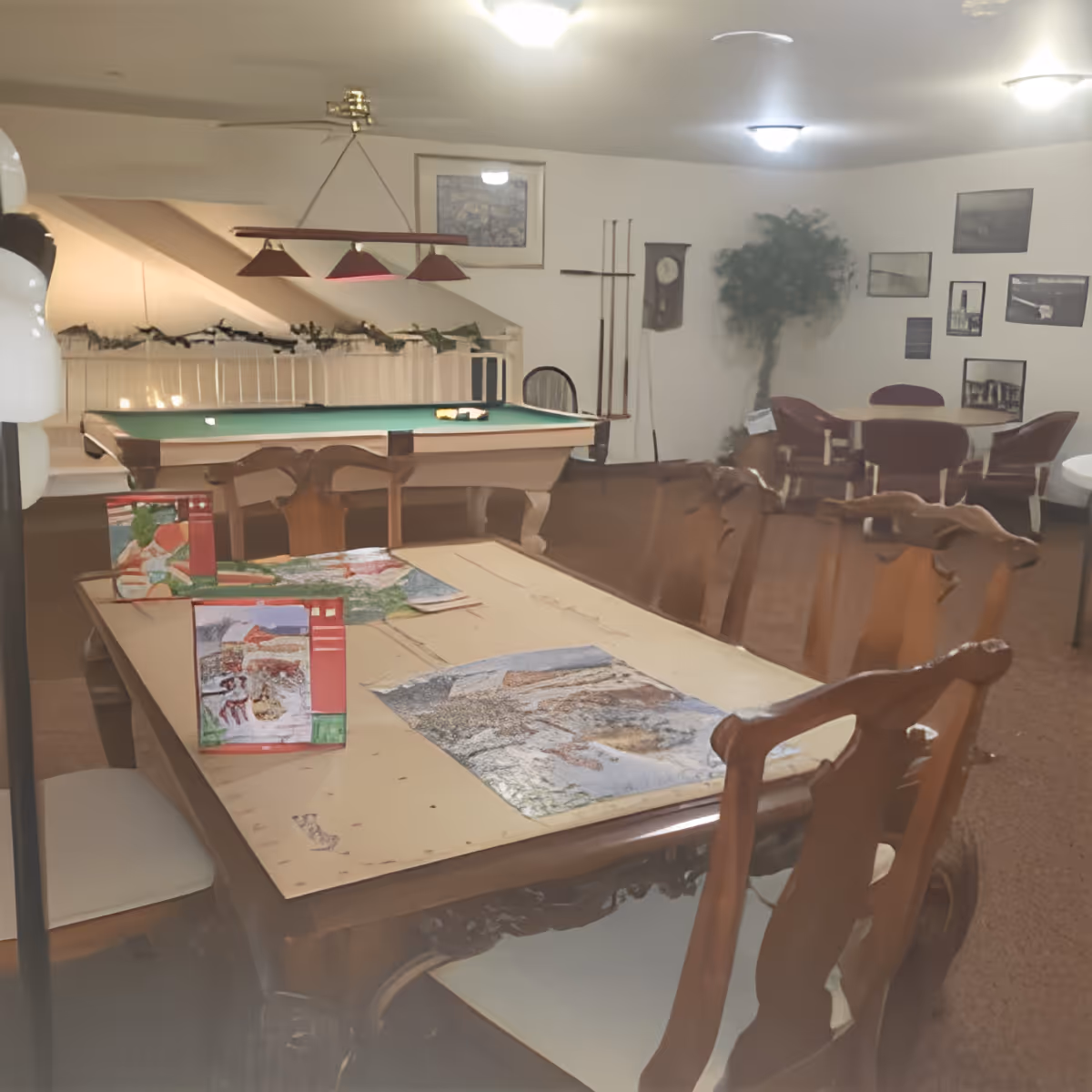 A communal interior room with a pool table, a long wooden table covered in puzzles and chairs, and additional seating and wall decor in the background.