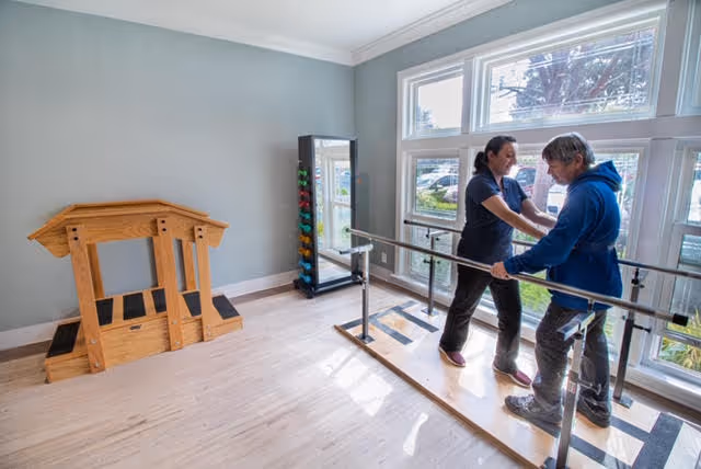 A therapy room with large windows letting in natural light. A caregiver assists an elderly person walking between parallel bars used for physical rehabilitation. There is a wooden step exercise structure and a rack with colorful weights against the wall.