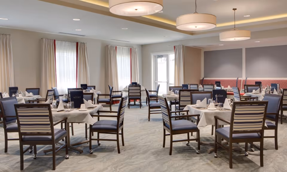 A spacious dining room with multiple tables covered in white tablecloths, each set with napkins, glasses, and menus. The room has large windows with curtains, modern ceiling lights, and comfortable chairs with striped backs and blue cushions.