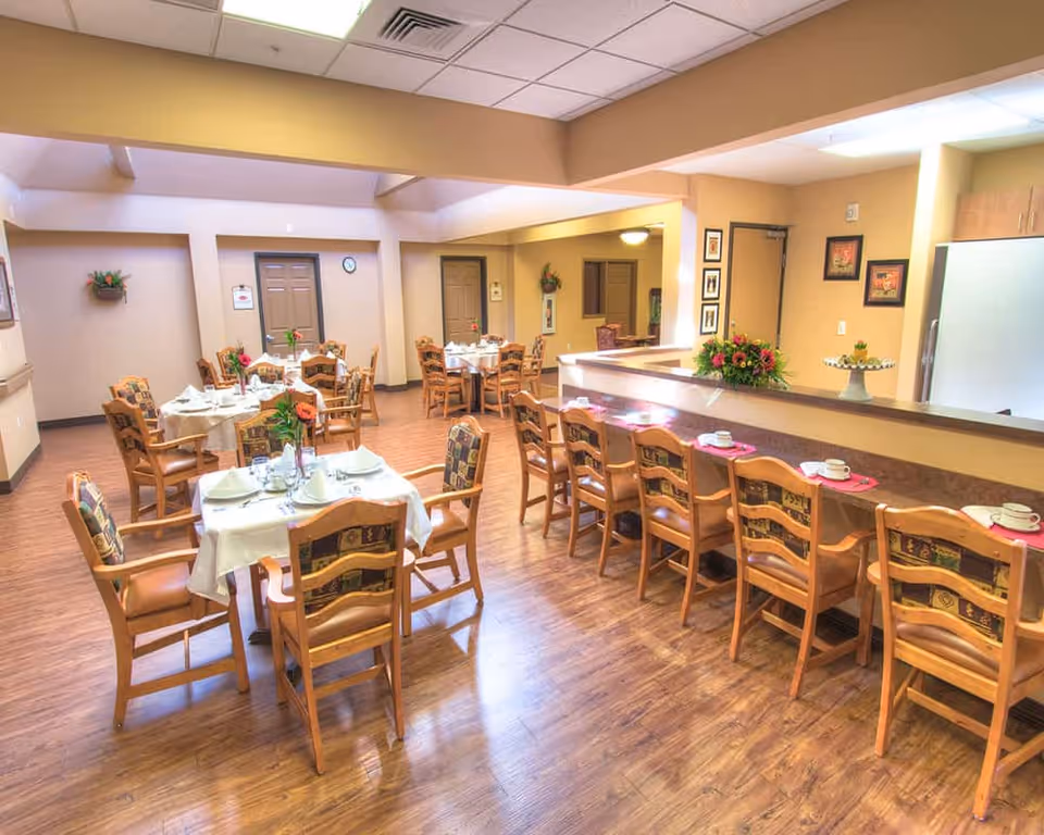A bright dining room in a senior living facility with several round tables covered with white tablecloths, each set with plates, cups, and floral centerpieces. There is a long counter with wooden chairs lined up, also set with cups and saucers. The room has wooden flooring, beige walls, and a ceiling with recessed lighting. Decorative framed pictures and plants adorn the walls.