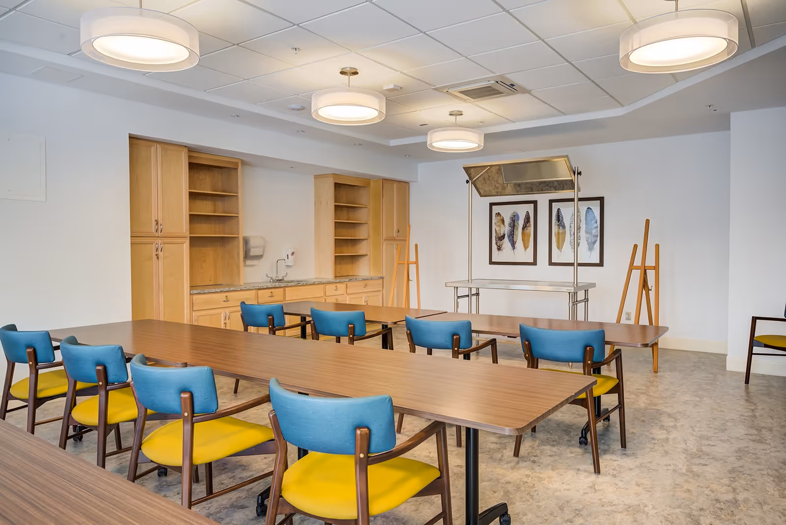 A bright room with long wooden tables and chairs with blue backs and yellow seats arranged in rows. The room has light wood cabinets with granite countertops, a sink, and three framed feather artworks on the wall. There are also two easels and a metal warming tray stand. The ceiling has round modern light fixtures.