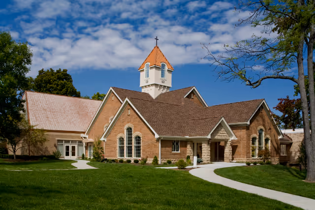 A brick building with a steep roof and a small tower topped with a cross, surrounded by green grass and trees under a partly cloudy blue sky.
