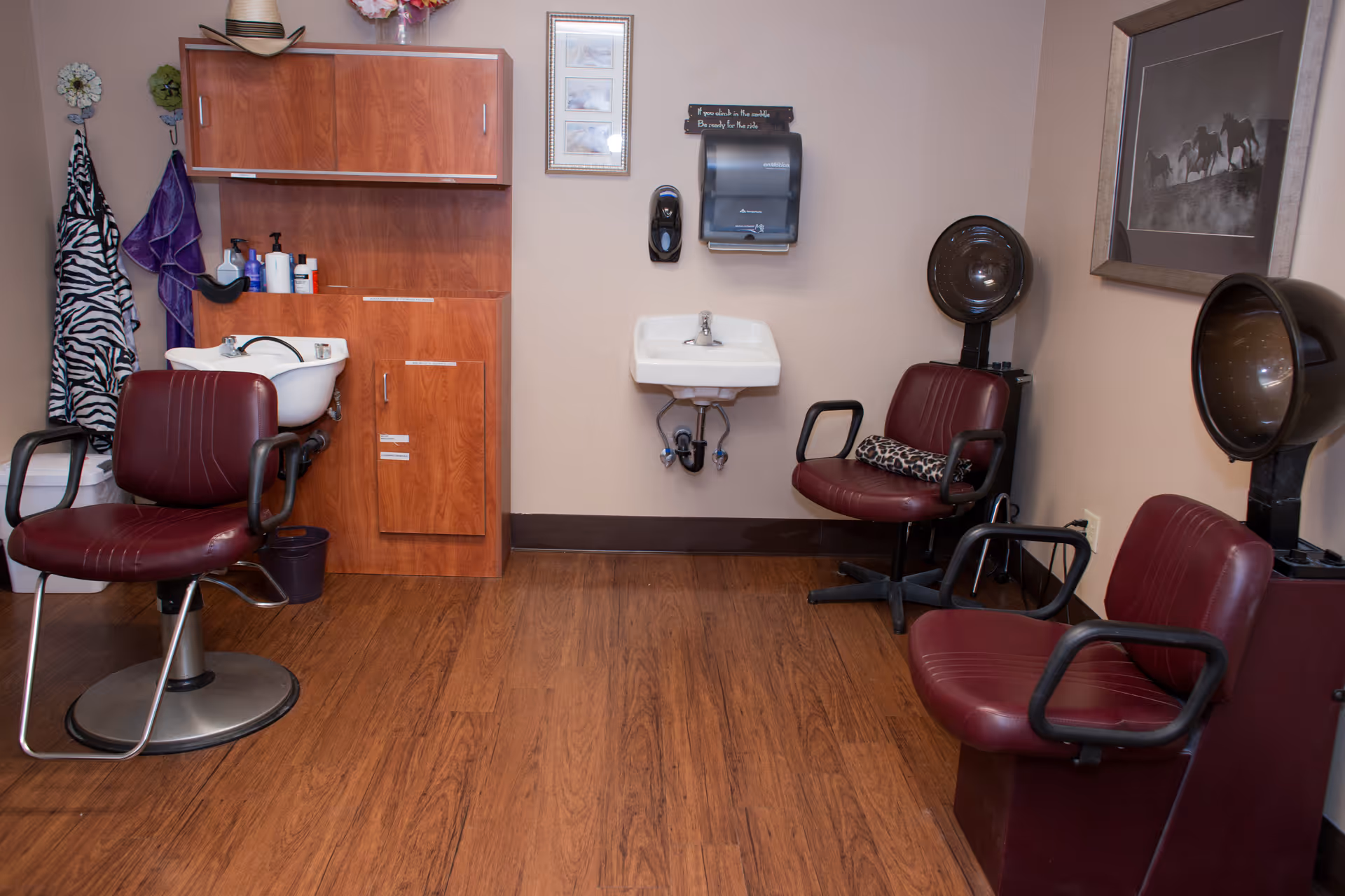 Small salon room with maroon styling chairs, a shampoo bowl, wall-mounted sink, and hooded hair dryers.