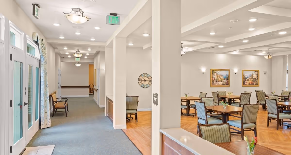 Interior view of a senior living facility showing a hallway with seating along the wall and an adjacent dining area with multiple tables and chairs. The space is well-lit with ceiling lights, decorated with framed artwork, and has a clean, welcoming atmosphere.