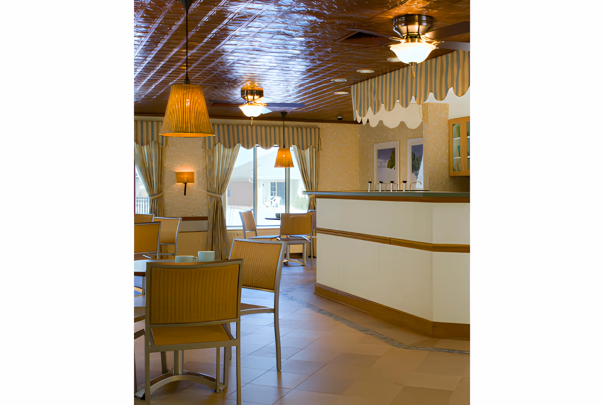 Dining room with wooden chairs and tables, hanging pendant lights, and a service counter beneath a decorative ceiling.