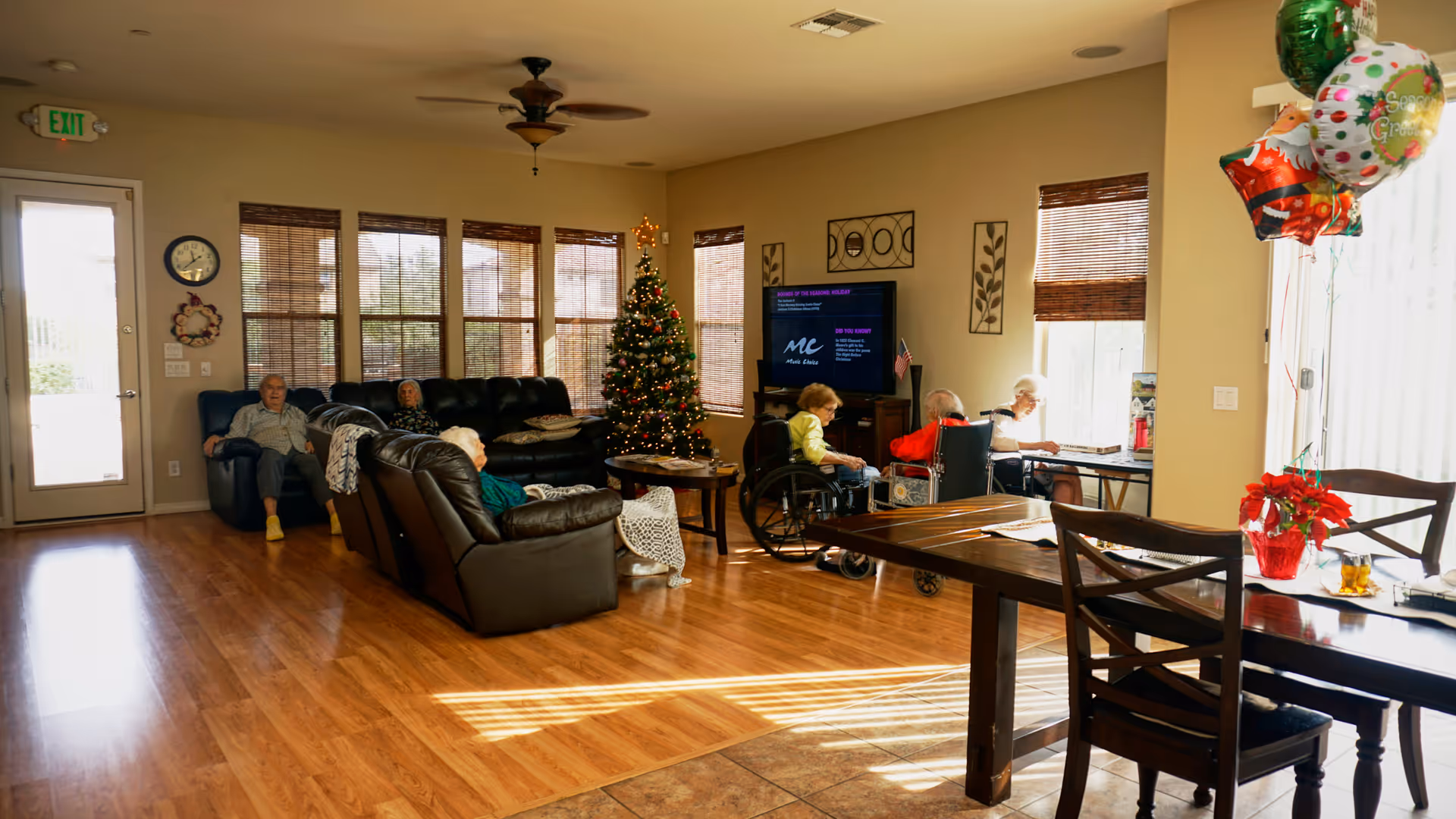 A bright living room in a senior living facility with several elderly residents seated on sofas and in wheelchairs. There is a decorated Christmas tree near the windows, a TV on a stand, and a dining table with chairs and festive balloons. The room has wooden floors and large windows with blinds letting in natural light.
