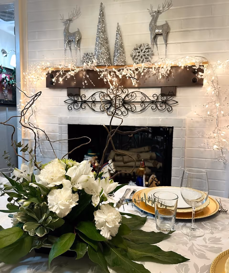 Festively decorated dining table with a white floral centerpiece in front of a lit, garland-adorned fireplace and mantel topped with silver holiday decorations.