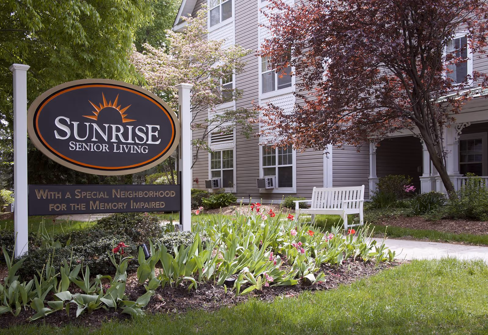 Outdoor view of Sunrise Senior Living facility with a sign that reads 'Sunrise Senior Living With a Special Neighborhood for the Memory Impaired' surrounded by green plants and flowers, a white bench, and a building with windows and trees in the background.