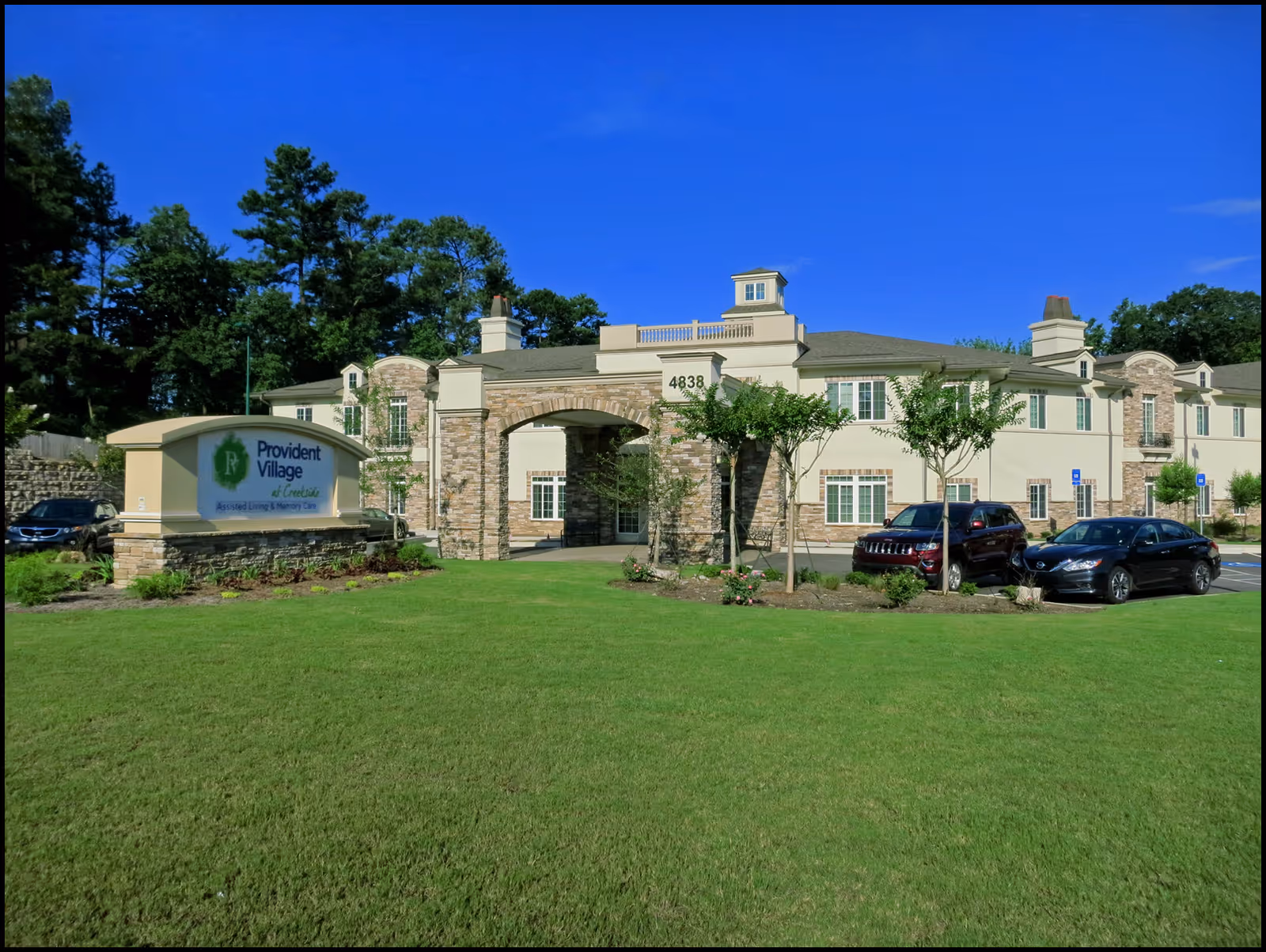 Exterior view of a large assisted living facility with a manicured lawn and parked cars.