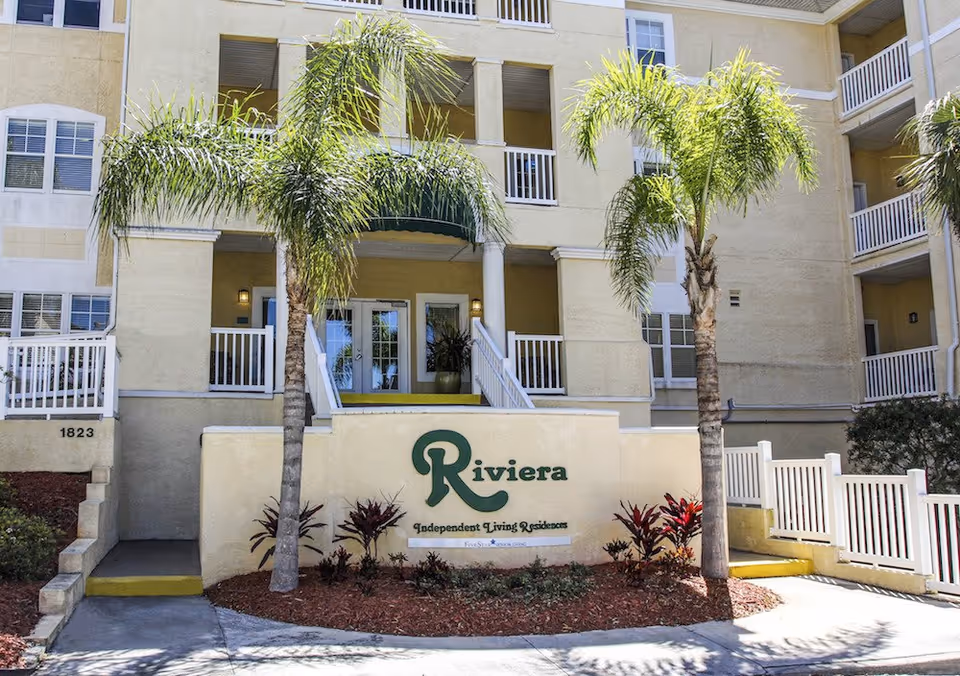 Front entrance of the Riviera senior living building showing the sign, palm trees, and balconies.