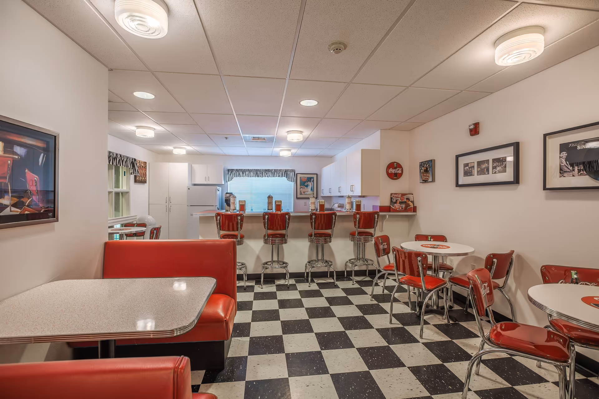 A retro-style dining area with red vinyl chairs and booths, black and white checkered floor, round white tables, and a counter with red bar stools. The walls are decorated with vintage Coca-Cola signs and framed pictures. The kitchen area is visible in the background with white cabinets and appliances.
