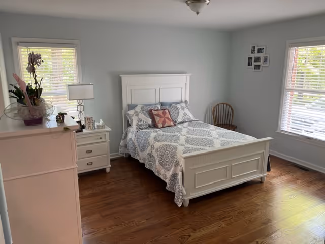 Light blue bedroom with white bed, dresser, nightstand, windows, and hardwood floor.