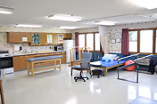Bright multipurpose therapy/activity room with a kitchenette, treatment table, exercise equipment, chairs, and large windows.