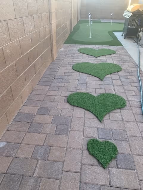 Narrow paved outdoor walkway with heart-shaped artificial turf pads leading to a small putting green beside a block wall and a grill.