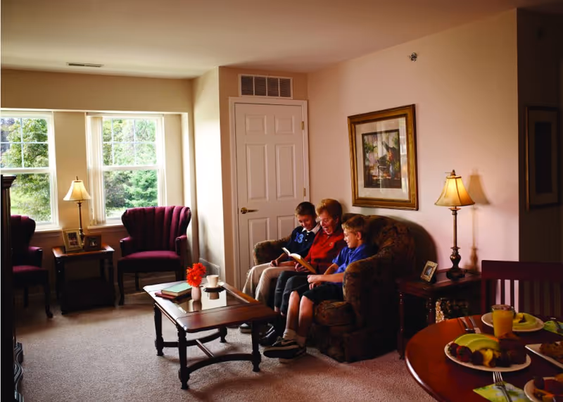 An inviting living room where an older woman sits on a couch reading with two boys, with chairs, lamps, a coffee table, and a dining table with plates in the foreground.