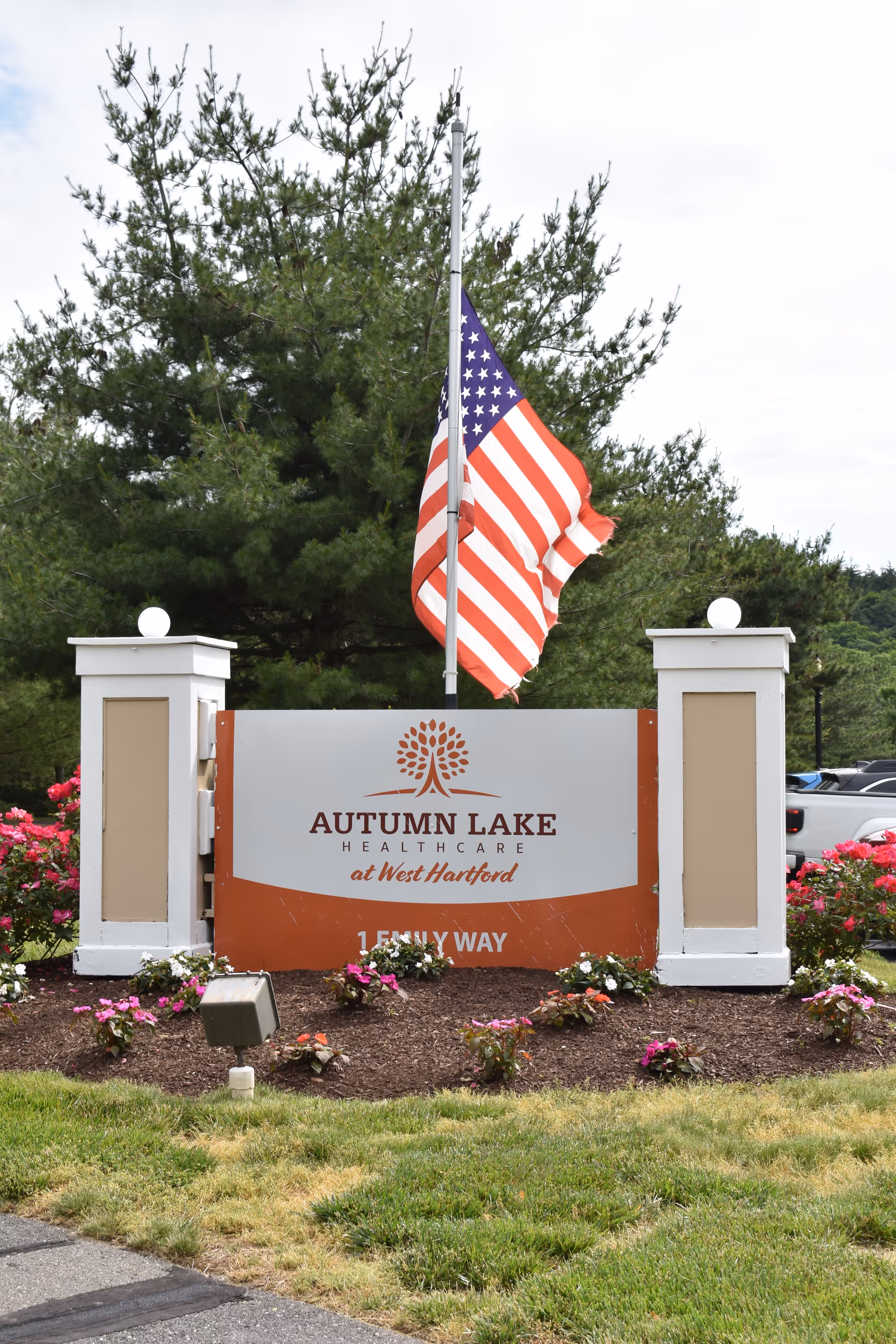 Outdoor sign for Autumn Lake Healthcare at West Hartford with an American flag flying at half-mast behind it, surrounded by flowers and greenery.