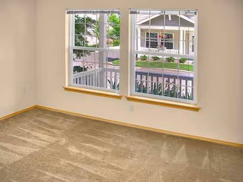 Empty beige-carpeted room with two windows overlooking a residential street.