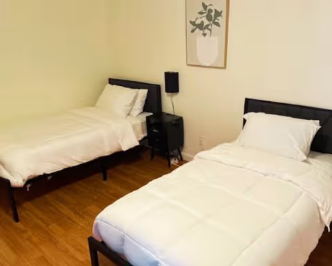 A simple bedroom with two single beds, each made with white bedding and pillows. Between the beds is a small black nightstand with a black lamp on it. A framed artwork featuring a plant hangs on the wall above the nightstand. The floor is wooden and the walls are painted light beige.