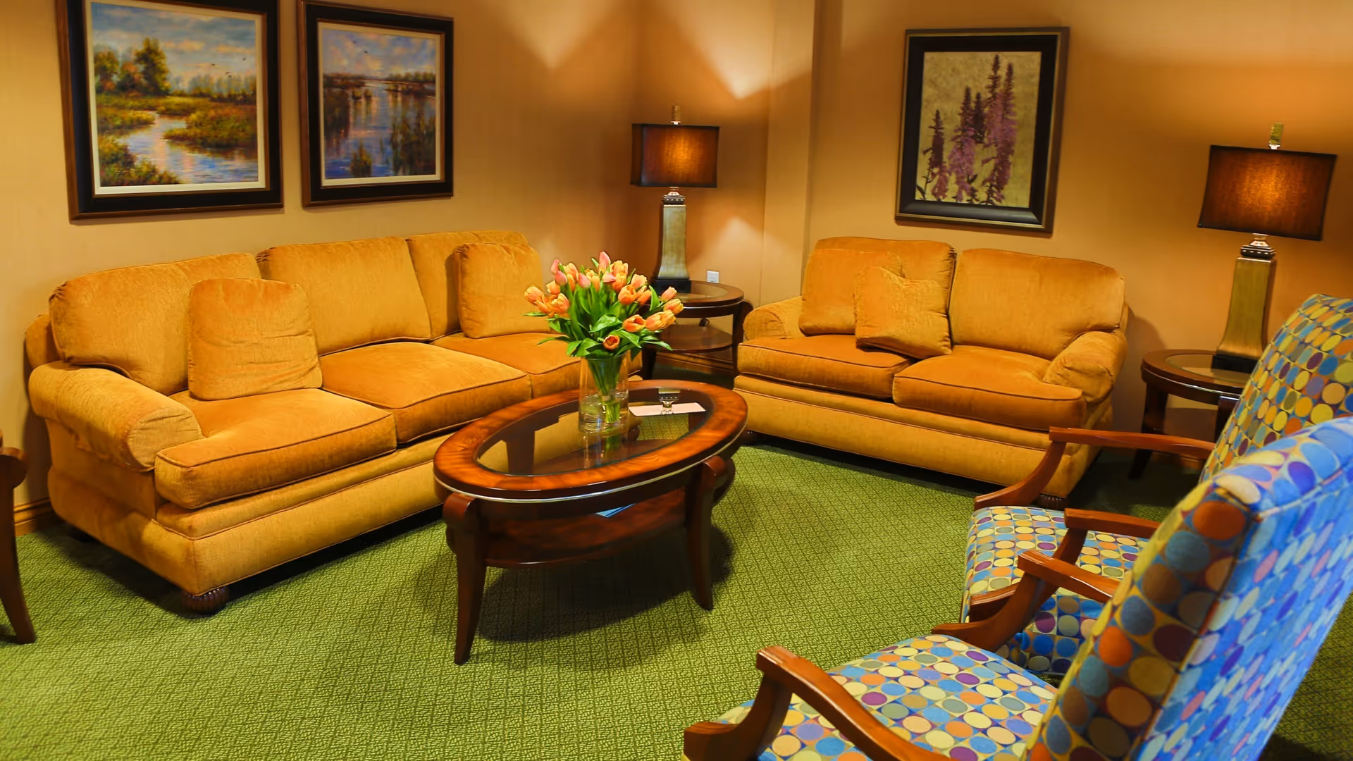 Warmly lit sitting area with two mustard-colored sofas, patterned armchairs, a wooden coffee table holding a vase of flowers, lamps, and framed artwork.