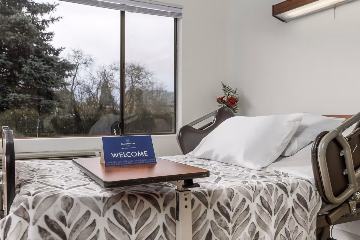 A hospital-style bed with white pillows and a patterned blanket in a room with a large window showing trees outside. A small table on wheels is positioned over the bed with a blue welcome sign on it. There is a small vase with red flowers on a shelf above the bed.