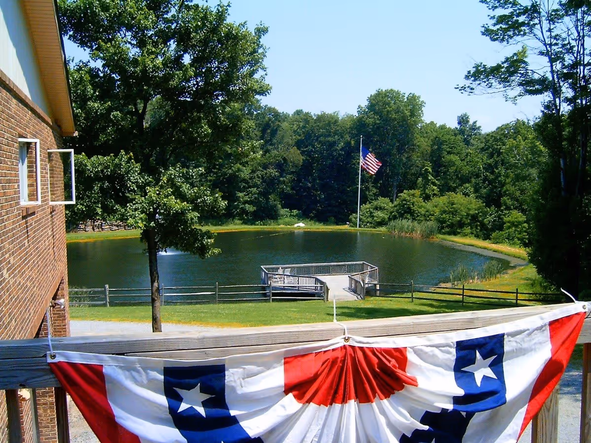View of a small pond with a wooden dock and an American flag on a flagpole near the edge of the pond, surrounded by green trees and grass. A brick building with an open window is visible on the left side, and a red, white, and blue patriotic banner is draped over a wooden railing in the foreground.
