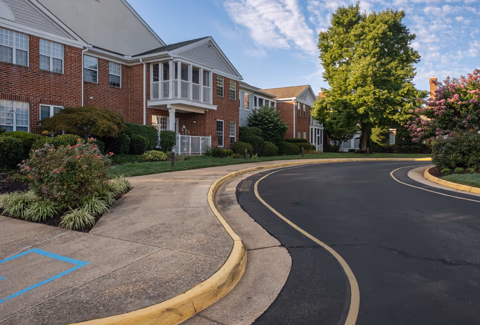 Curved driveway and sidewalk leading to a two-story brick building with white trim and multiple windows, surrounded by well-maintained bushes, trees, and flowering plants under a partly cloudy sky.