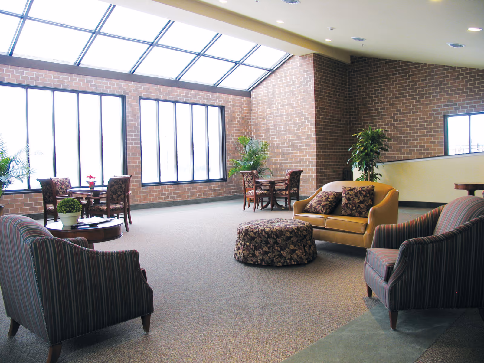 A bright and spacious common area with large windows and a skylight letting in natural light. The room features brick walls, carpeted floors, two striped armchairs, a mustard yellow loveseat with floral cushions, a floral patterned ottoman, and small round tables with chairs. There are also several potted plants adding greenery to the space.