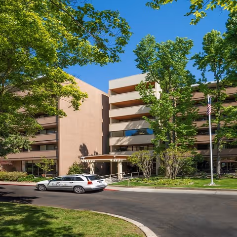 Front entrance of a multi-story senior living building with a driveway, parked vehicle, trees, and an American flag.