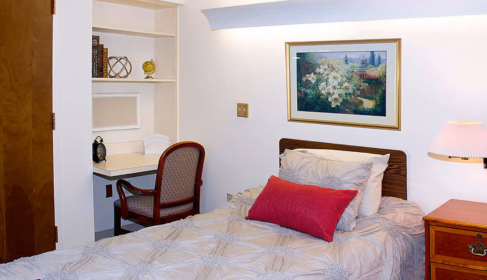 A cozy bedroom with a single bed featuring a beige comforter and a red accent pillow. Next to the bed is a wooden nightstand with a lamp. On the wall above the bed is a framed painting of white flowers in a garden. To the left, there is a built-in desk with a chair, some books, a decorative globe, and an alarm clock.