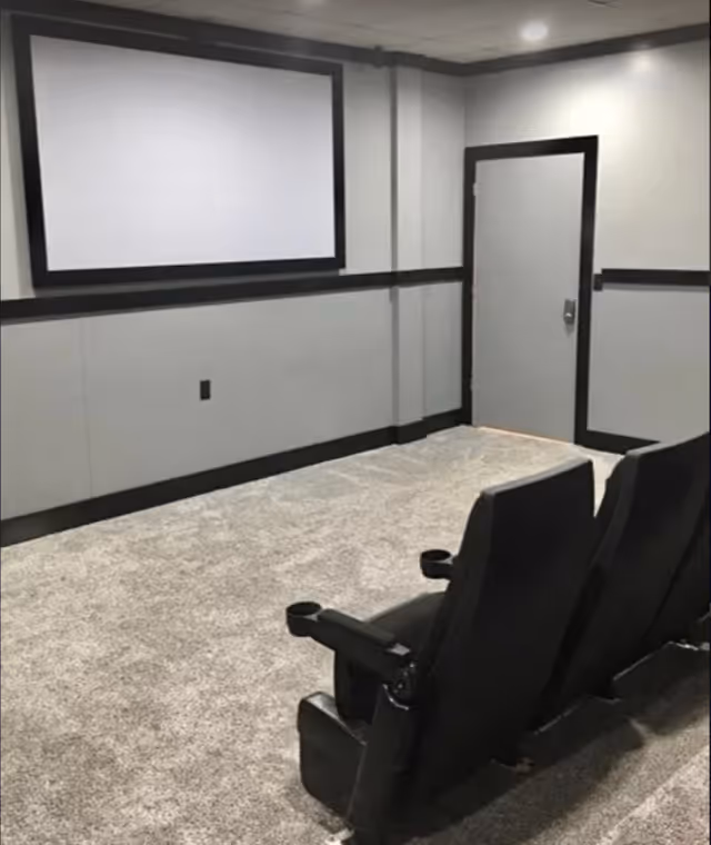 A small home theater room with two black reclining chairs facing a large blank projection screen mounted on a gray wall. The room has gray carpet, gray walls with black trim, a closed door, and recessed ceiling lights.