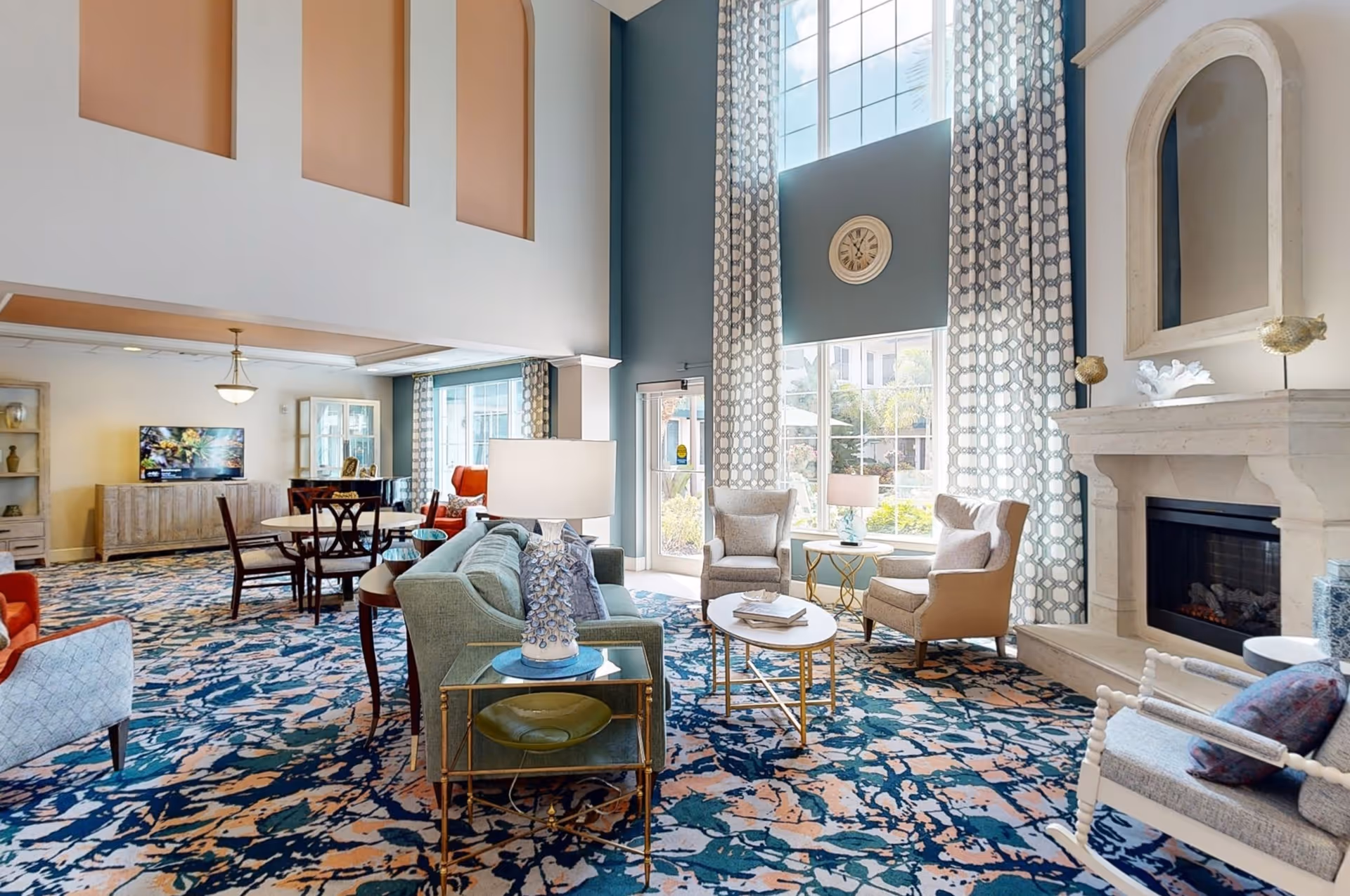 A spacious and well-lit living room area with high ceilings, large windows with patterned curtains, a fireplace, and comfortable seating including armchairs and sofas arranged around coffee tables. There is a patterned carpet covering the floor and a clock mounted on the wall above the windows. In the background, there is a dining area with a table and chairs and a TV on a cabinet.