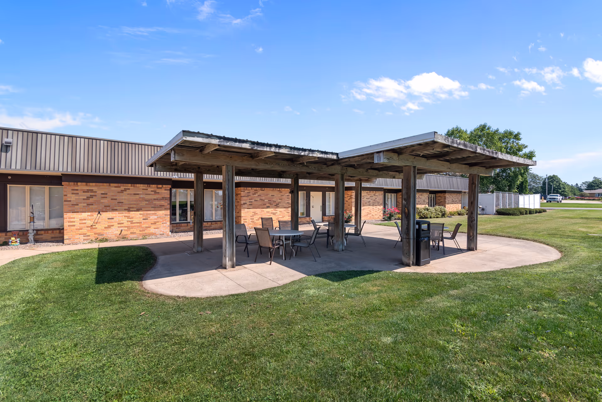 Outdoor covered patio area with several tables and chairs on a concrete slab surrounded by green grass, adjacent to a single-story brick building under a blue sky with some clouds.