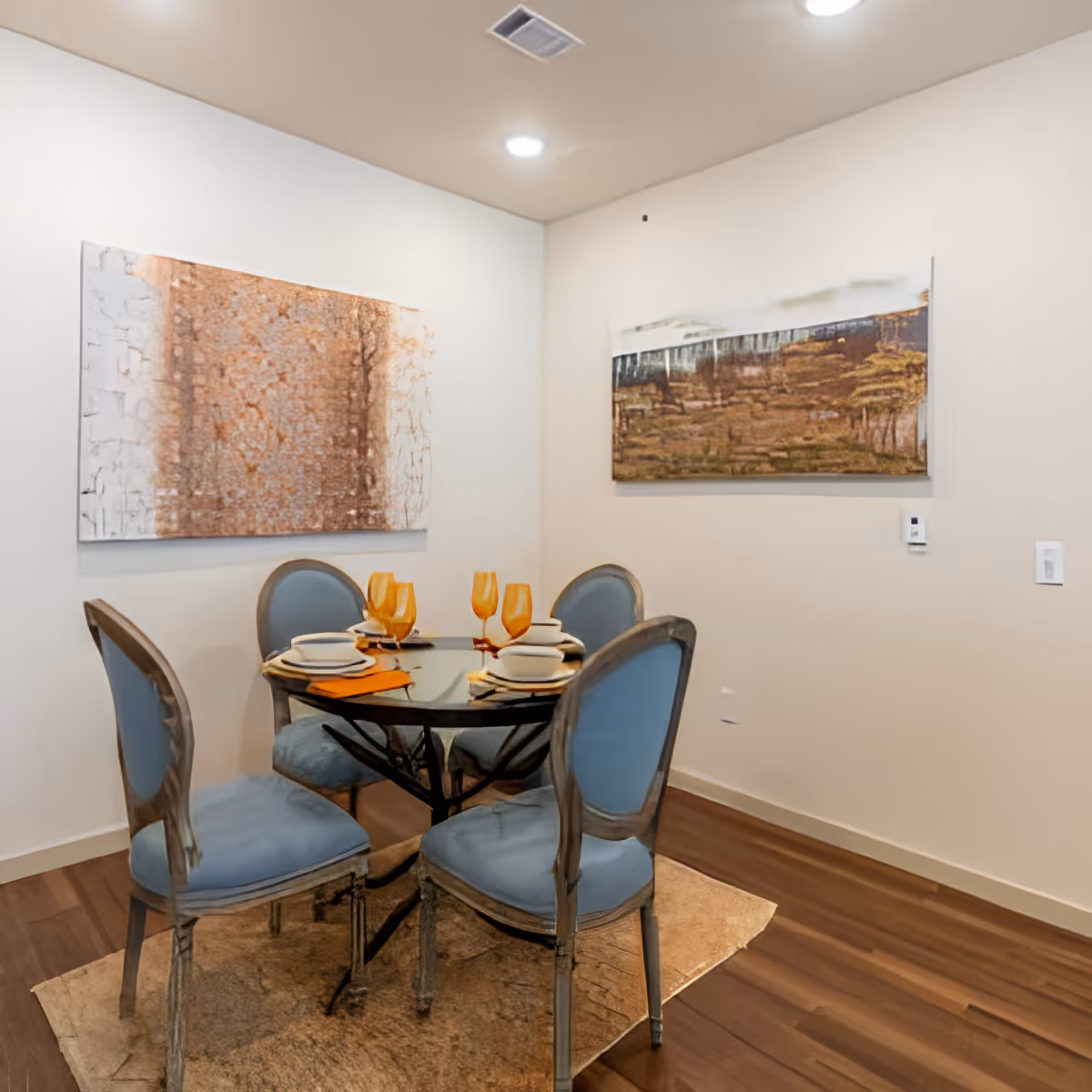 A small dining area with a round glass table set for four, featuring blue cushioned chairs and orange-tinted glassware. Two abstract paintings hang on the beige walls, and the floor is wooden with a light-colored rug under the table.