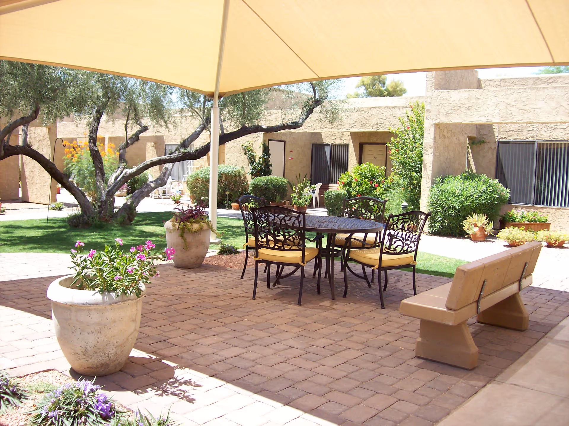 Outdoor patio area with a large beige canopy providing shade over a round metal table with four chairs that have yellow cushions. There is a beige bench nearby, large potted plants with flowers, green grass, and trees in the background. The surrounding building has a stucco exterior with windows and doors visible.