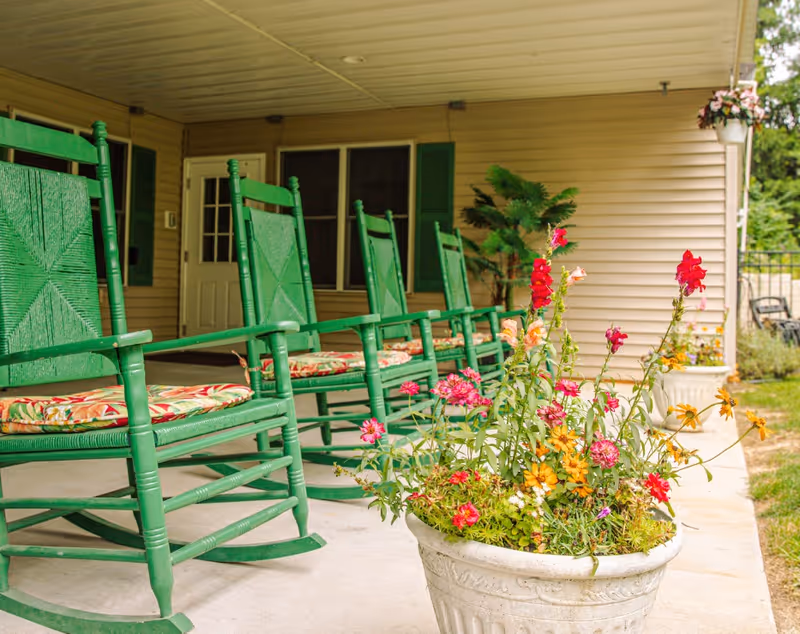 A row of green wooden rocking chairs with floral cushions on a covered porch. In the foreground, there is a large white planter filled with colorful flowers. The porch is attached to a beige building with green shutters and a white door.