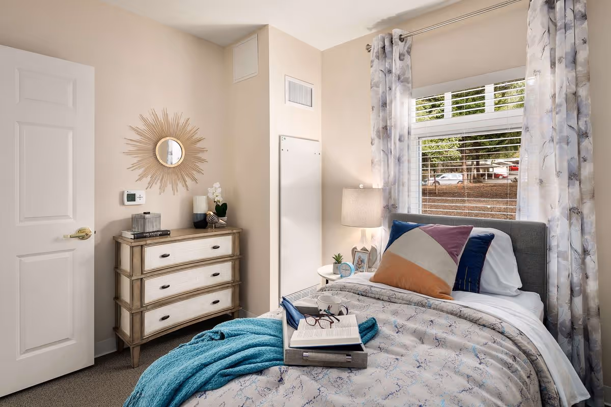 A neatly furnished small bedroom with a single bed, decorative pillows and throw, dresser with mirror, bedside table and a curtained window.