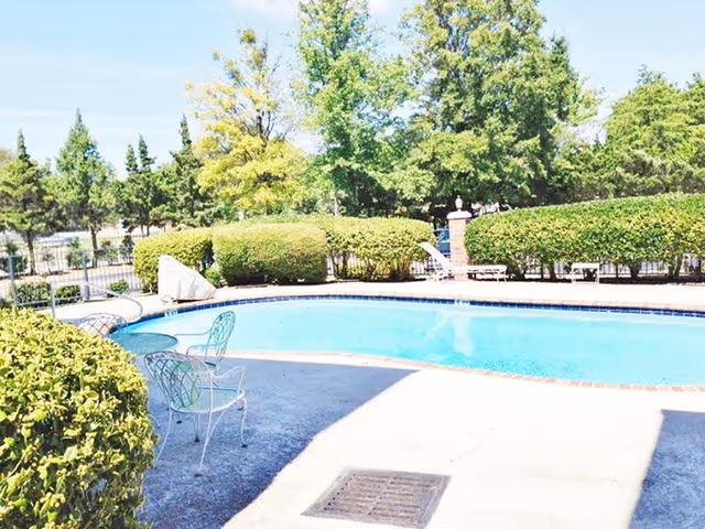 Outdoor swimming pool area surrounded by trimmed bushes and trees with two white metal chairs on the concrete pool deck.
