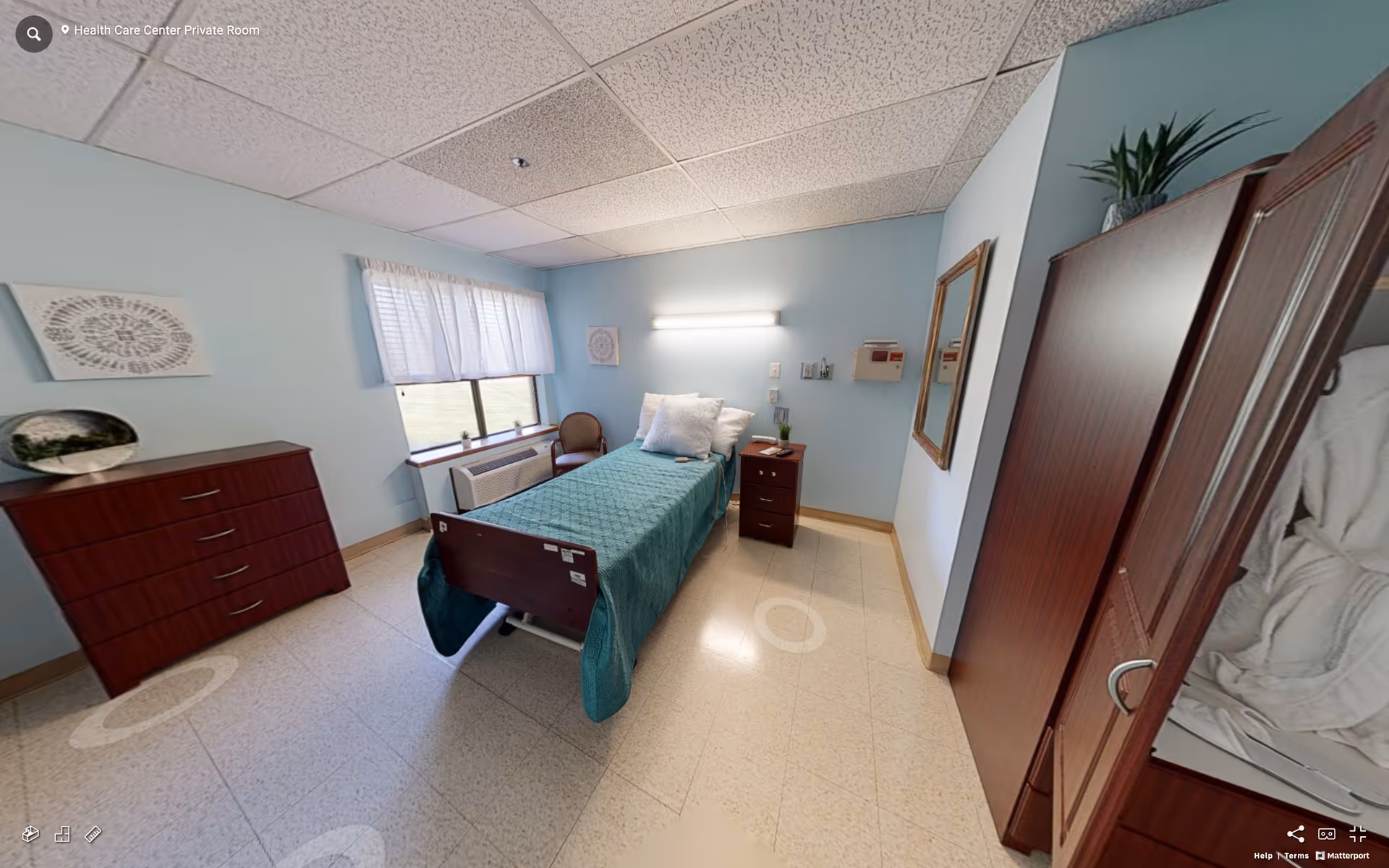 A private room in a health care center with a single bed covered in a teal blanket and white pillows. The room has light blue walls, a window with white curtains, a wooden dresser, a small wooden nightstand with a plant and a lamp, a wooden chair near the window, and a wooden wardrobe with a plant on top. The floor is tiled, and there is a large mirror on one wall.