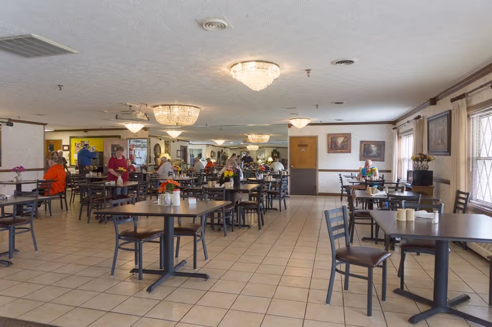Spacious dining room with multiple tables and chairs, chandeliers overhead, and several residents seated and standing throughout the room.