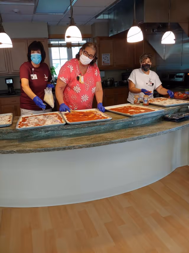 Three women wearing masks and gloves preparing multiple rectangular pizzas on trays in a kitchen with wooden cabinets and hanging lights above the counter.